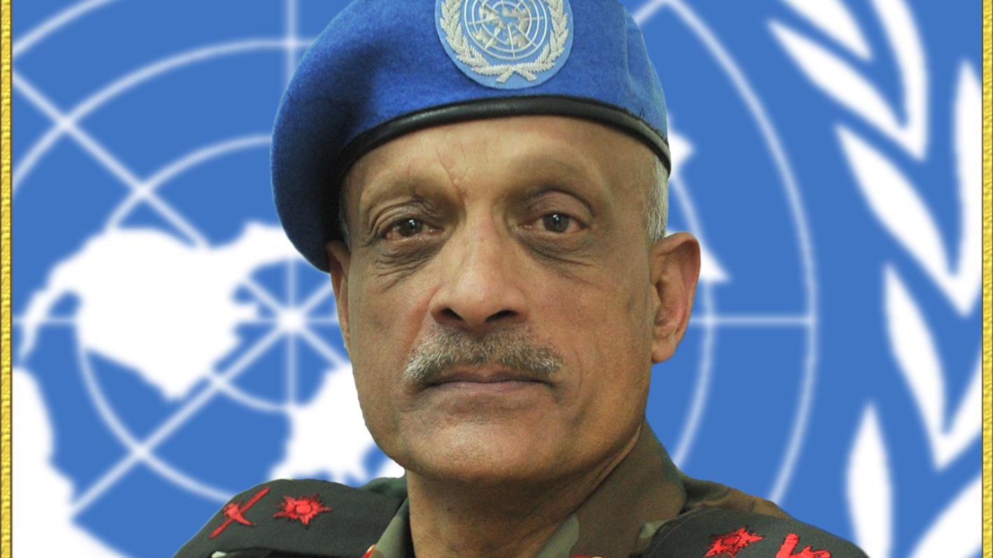 A man in military uniform with a blue beret sits in front of the UN logo